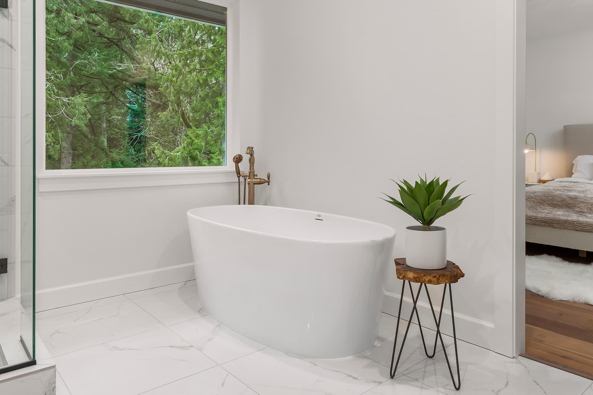Freestanding white bathtub beside a window in a bathroom with marble-look floor tile